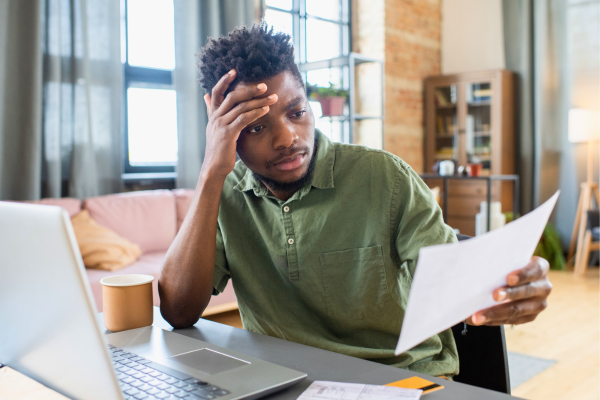 A stressed person working on laptop holding a paper.