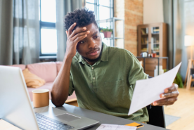 A stressed person working on laptop holding a paper.