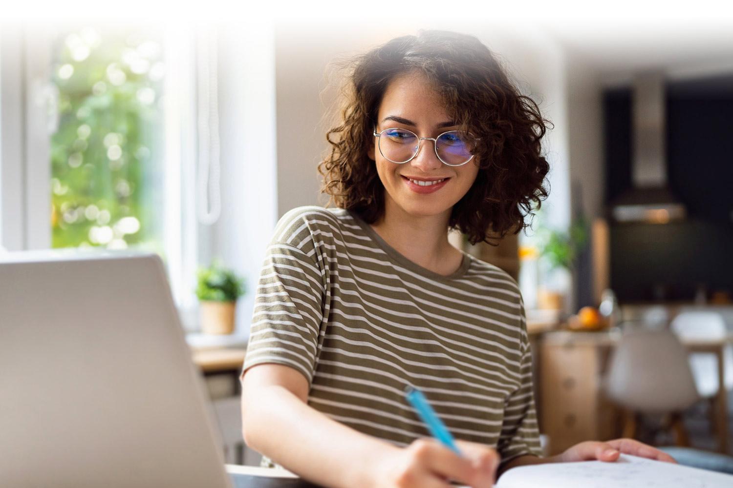 Young hispanic woman looking at the computer and taking notes at her desk at home