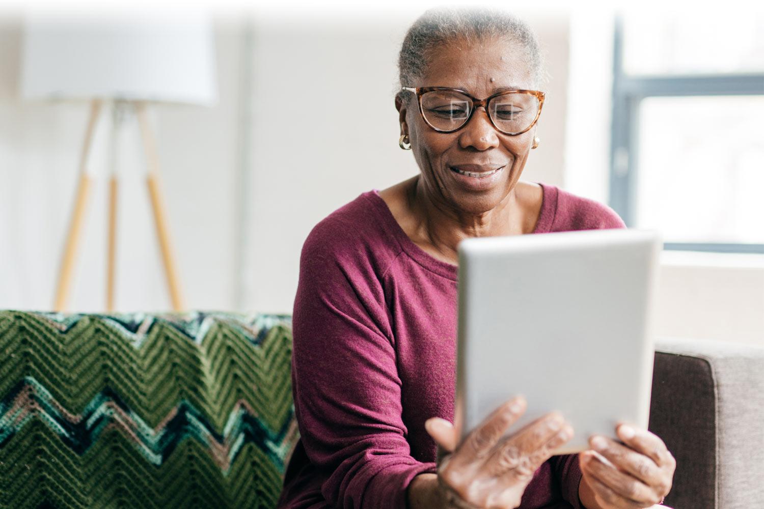 Older woman sitting on the couch looking at a webpage on her tablet.