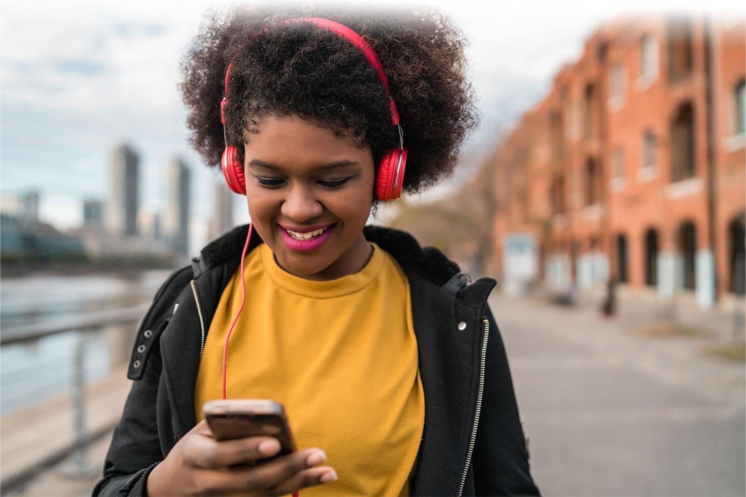 Woman listening to headphones and looking at phone while standing on the sidewalk