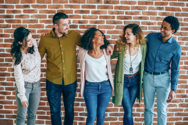 Five smiling people stand close together against a red brick wall, dressed in casual attire.